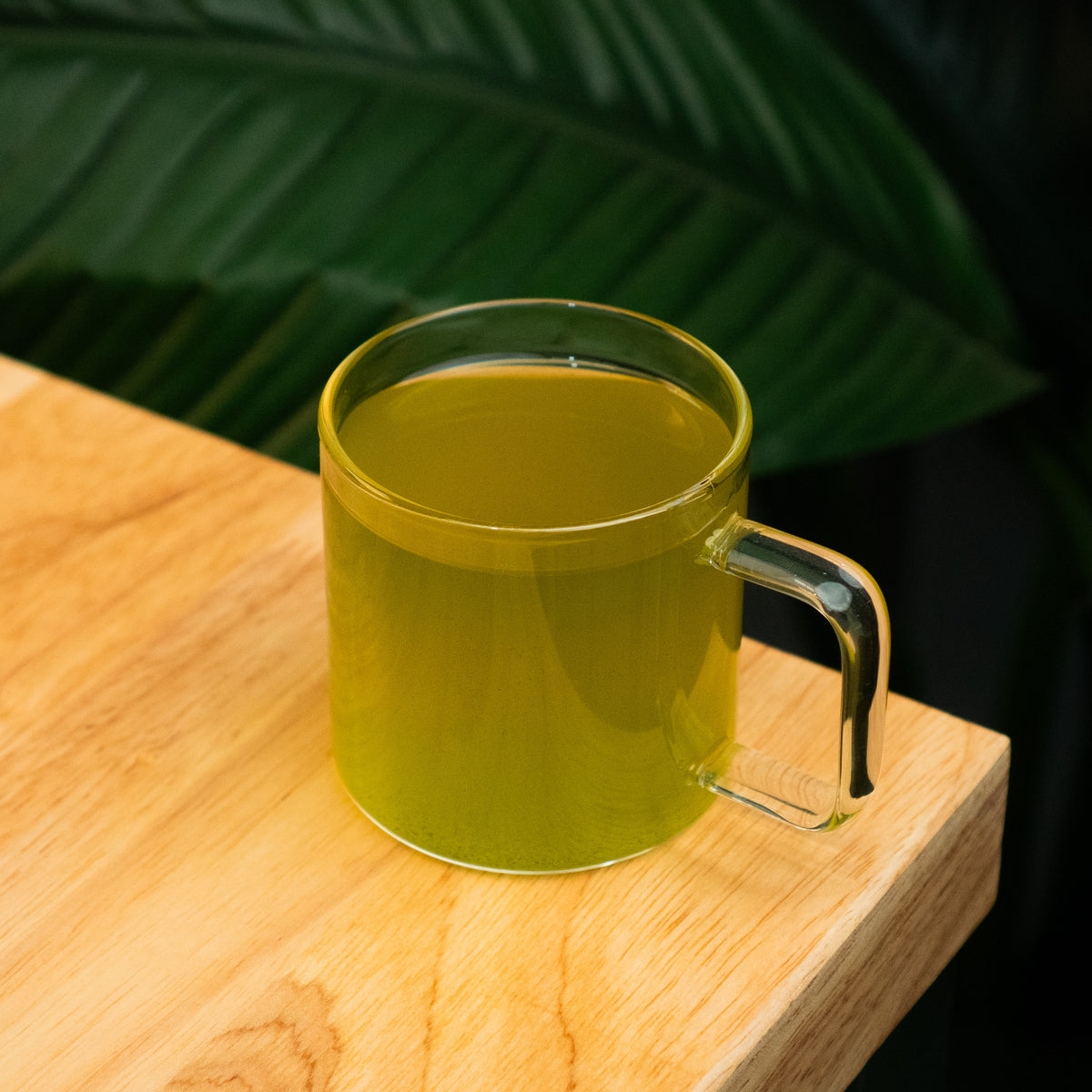 Clear mug with green tea on a wooden surface with leafy background