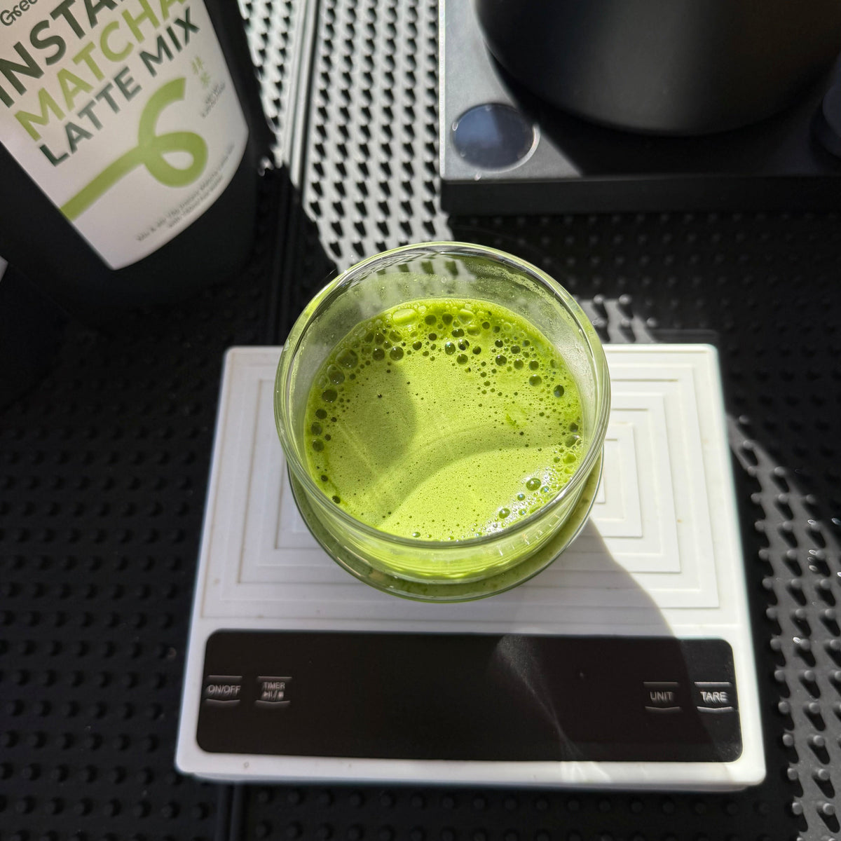 Green matcha latte mix in a glass on a digital scale with a package of matcha latte mix in the background.