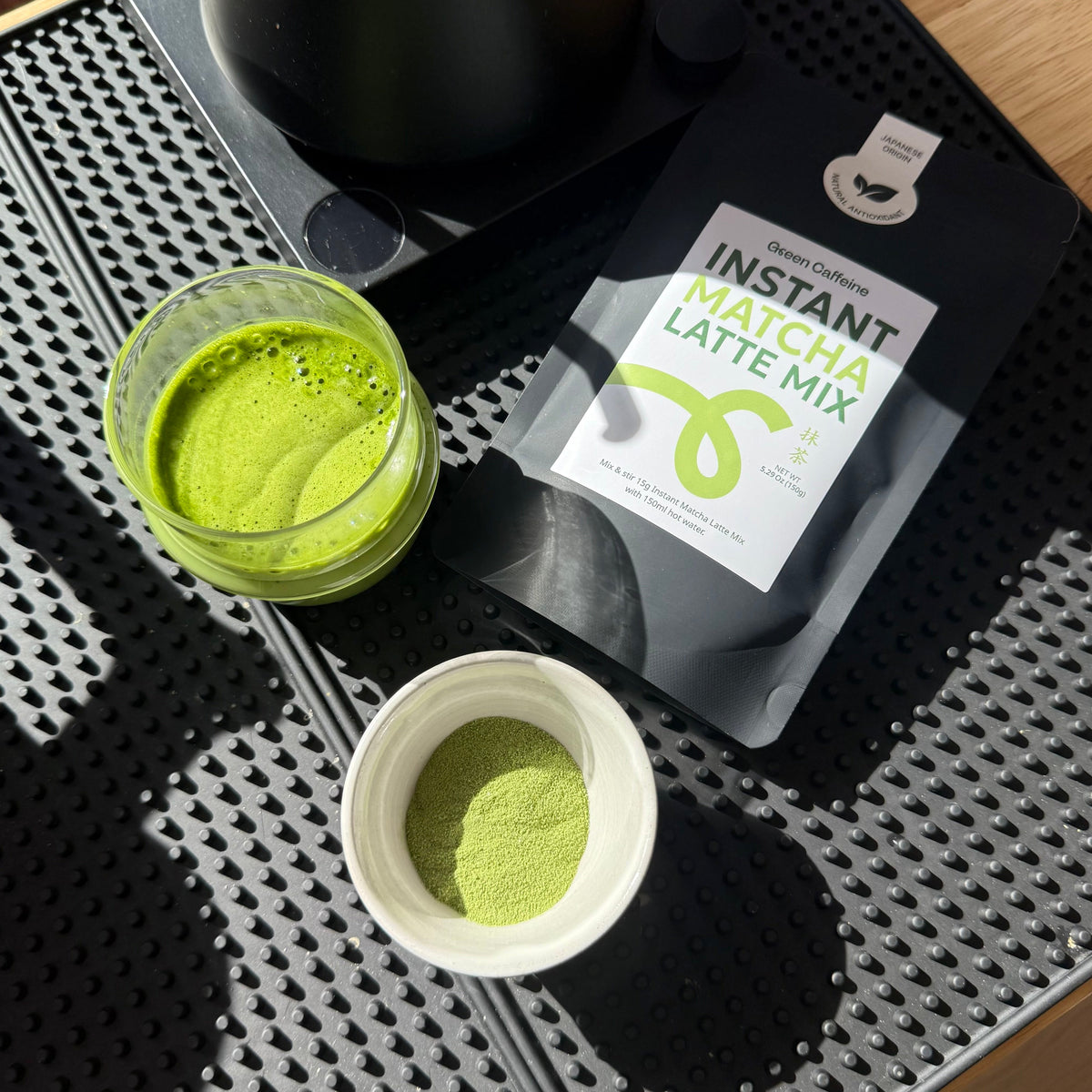 Matcha latte mix packaging with a container of matcha powder on a black textured mat.