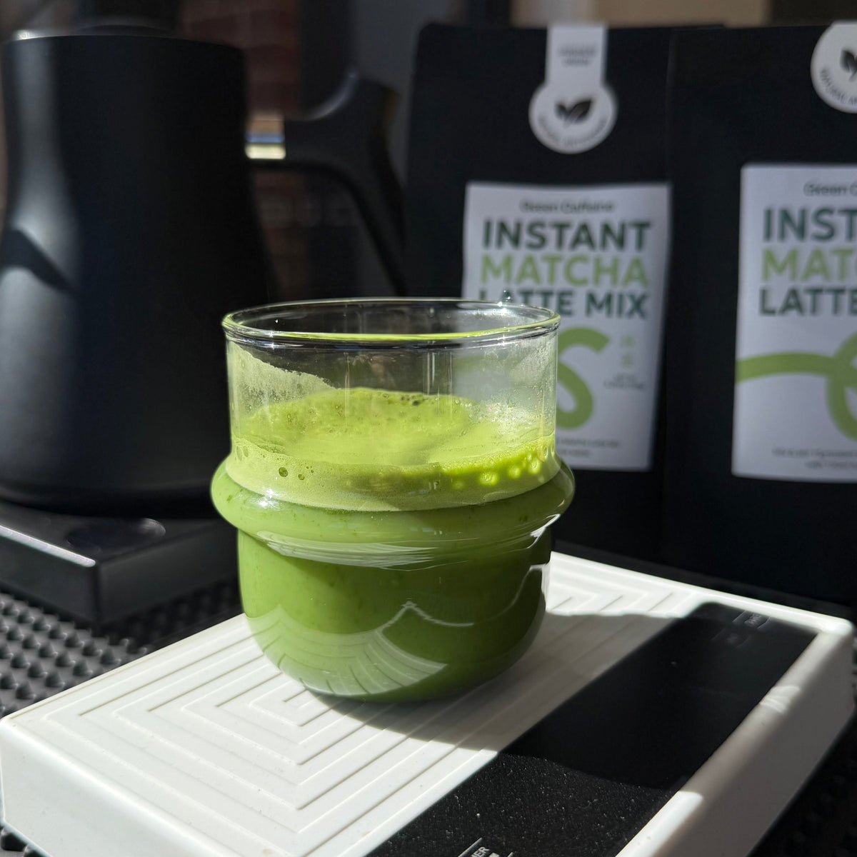 Green matcha latte in a glass on a digital scale with matcha latte mix packets in the background.